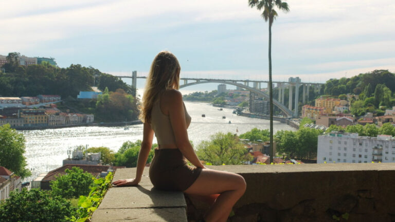 Author Sky Ariella sitting on a stone ledge overlooking the Douro River with the Arrábida Bridge in the background