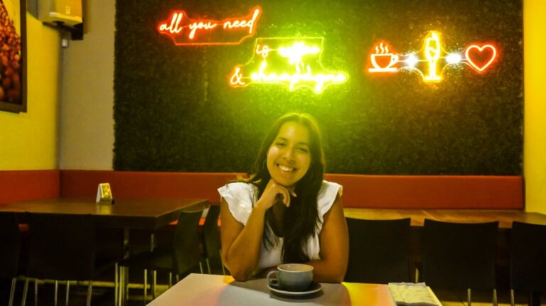 The author Vanessa Ramos, posing for a photo with a cup of coffee and neon lights in the background at Café Lucero