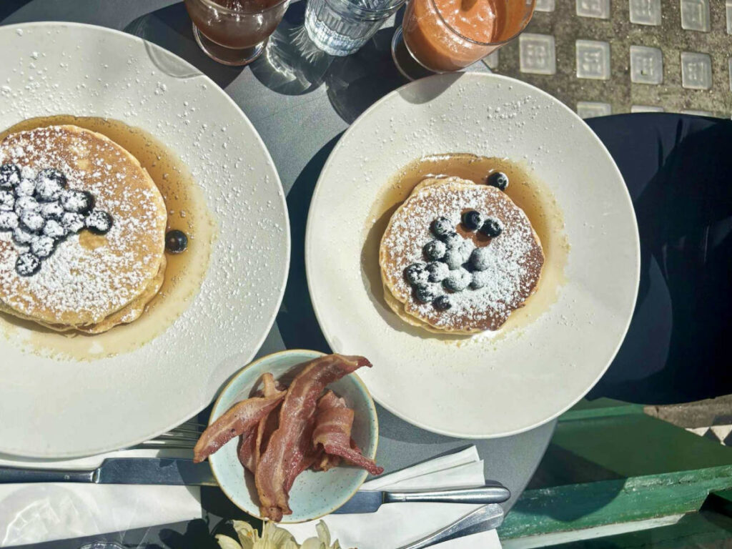 Two plates of blueberry pancakes with maple syrup and a side of bacon