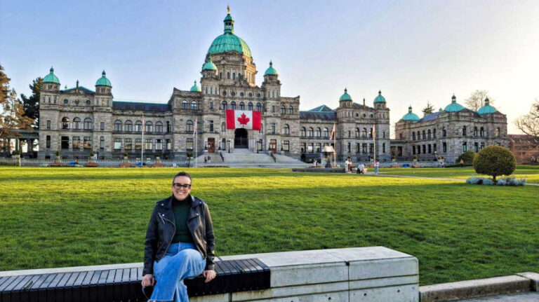 The author Catherine Driver sitting in front of the BC’s Parliament Buildings with a flag of Canada