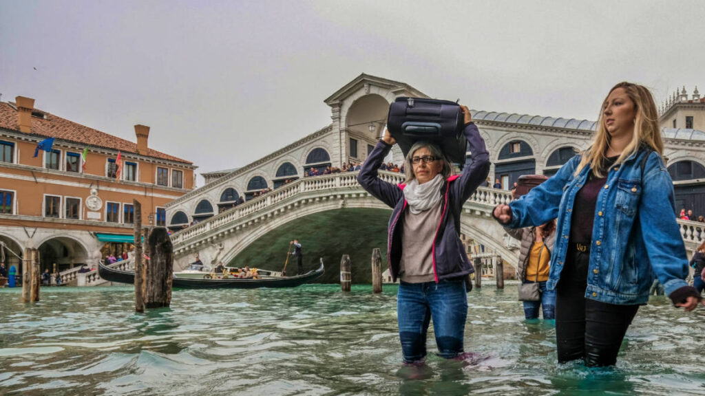 The tourists walking on the flooded streets of Venice while carrying their luggage, Climate Change Could Ruin These Tourism Destinations