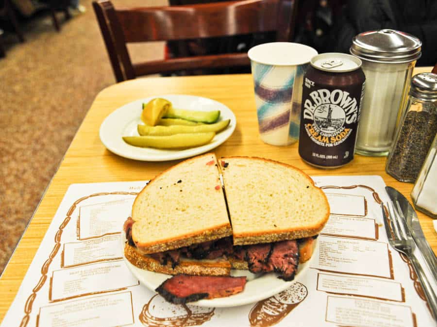 A plate of huge sandwich with pickles and drinks on the side from Katz Deli